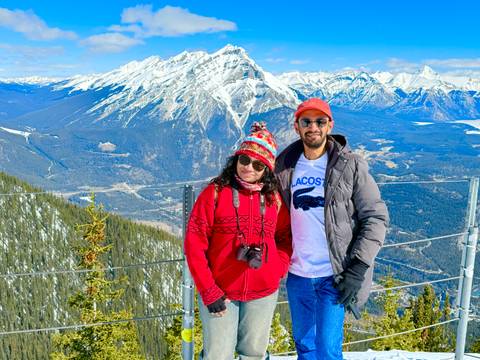       A couple poses at a fenced mountain viewpoint high above a winding river valley and snow-peaked ranges.
  
