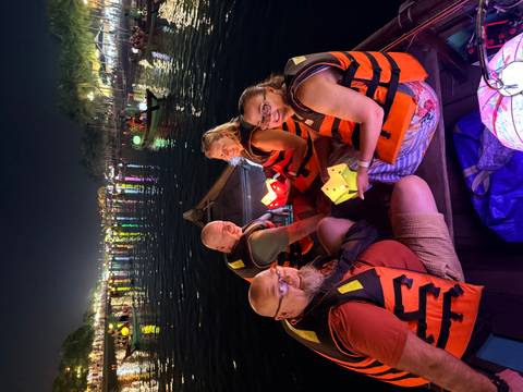       Four tourists in life vests seated in a wooden boat releasing paper lanterns onto a river at night
  