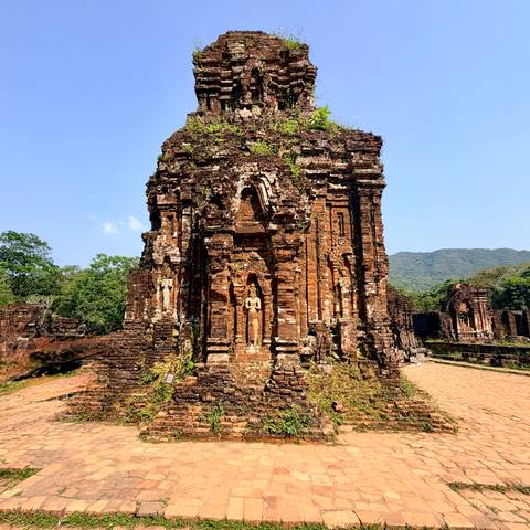       Ancient red-brick Hindu temple ruins surrounded by greenery and distant mountains under clear skies
  