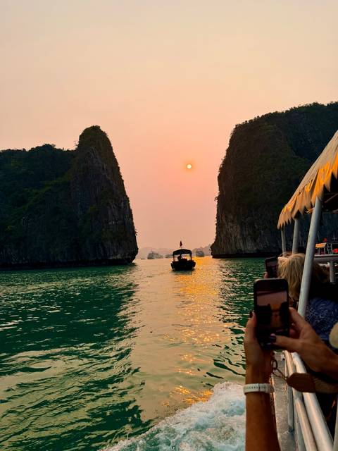       Boat heading between towering limestone cliffs at orange sunset reflecting on emerald water
  