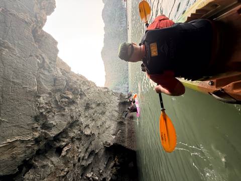       Kayaker paddling from inside a limestone cave toward open bay waters with cliffs beyond
  