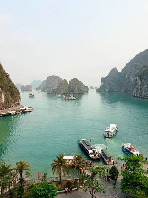       Panoramic view of turquoise bay dotted with boats and limestone islands under hazy sky
  