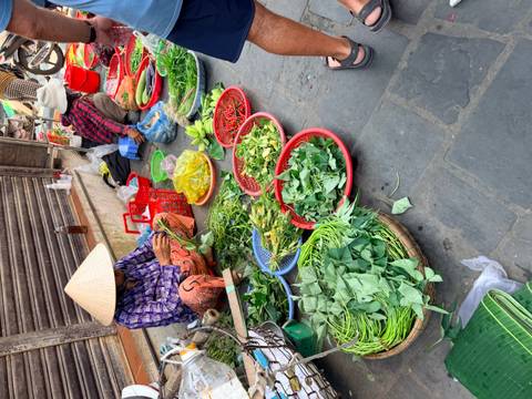       Local market stall with vendor in conical hat selling baskets of fresh green vegetables and chilies
  