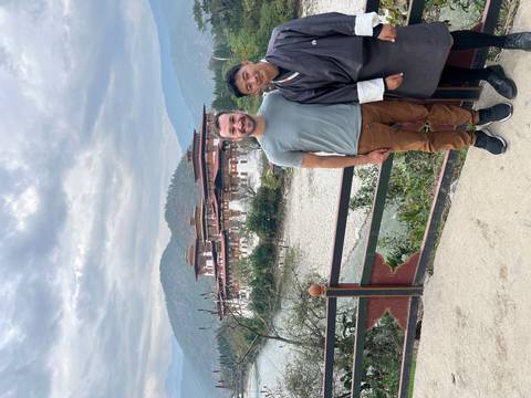       Two travellers posing in front of Punakha Dzong beside river under cloudy sky
  