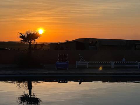       Sun setting behind a desert camp and palm tree, reflected in a swimming pool at the foreground.
  