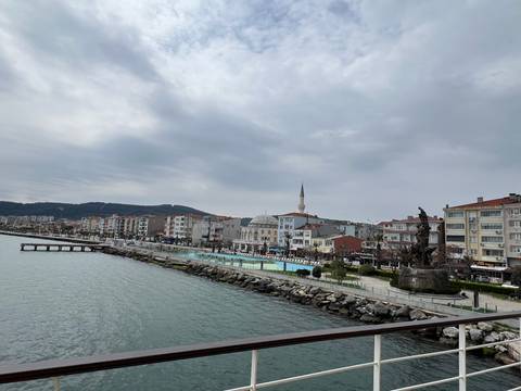       Seaside Turkish town with mosque, waterfront promenade and distant wooded hills under gray skies.
  