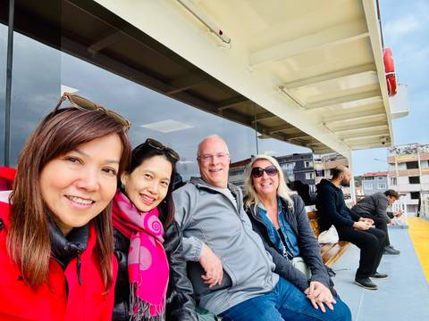       Smiling travelers seated on the deck of a ferry with a coastal town visible behind glass windows.
  