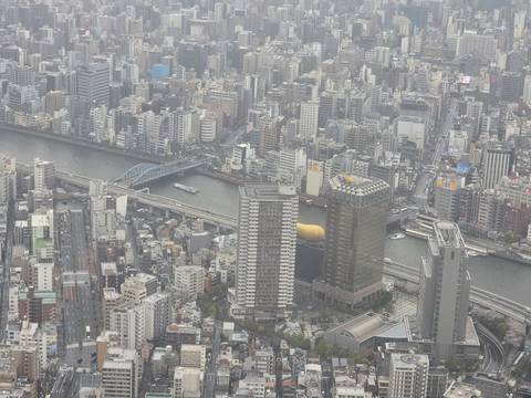       Aerial cityscape view over dense Tokyo buildings and the Sumida River with bridges and boats.
  
