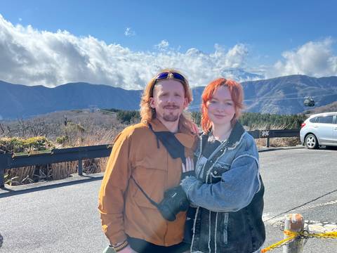       Young couple posing on mountain viewpoint with clouds, cable car and parked car behind.
  