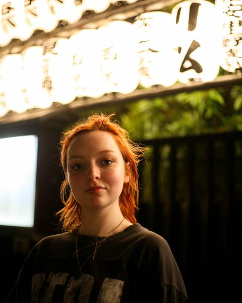       Close portrait of red-haired woman with soft background of greenery and roof edge.
  