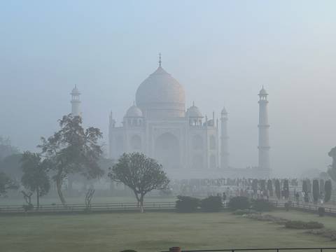       Taj Mahal shrouded in thick morning fog with silhouettes of visitors and trees.
  