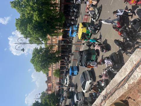       Busy Jaipur street scene with tuk-tuks, motorbikes and shoppers beneath pink sandstone facades.
  