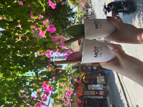       Two hands toast with signature herbal drinks in branded cups beneath bright bougainvillea on a sunny Hoi An street.
  