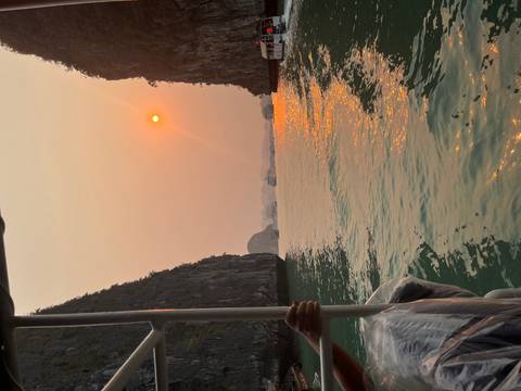       Sun setting through hazy sky over limestone karsts and emerald waters of Ha Long Bay as boats cruise between cliffs.
  