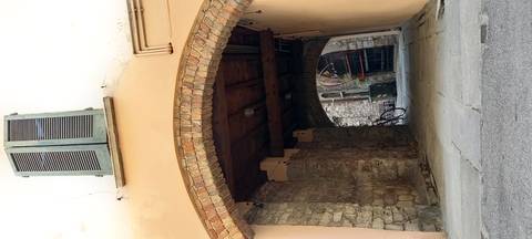       Stone archway leading to a quiet narrow alley with rustic walls, shuttered window and a parked bicycle at the far end.
  