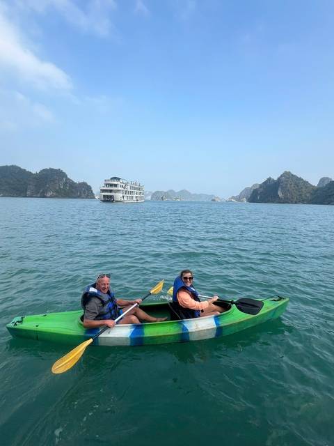       Two travellers in a green kayak paddle through the emerald waters of a karst-lined bay while a white cruise ship floats behind them.
  