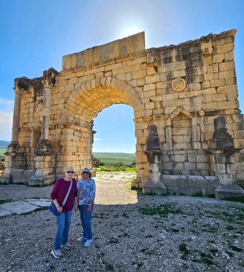       Two women posing before a large Roman stone arch with wide rural plains beyond.
  