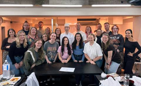       Large tour group poses with a sushi chef after a cooking class in a bright studio.
  