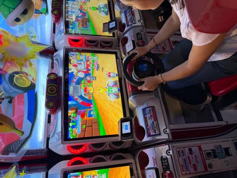       Player steers a Mario Kart arcade machine inside a colorful game center.
  