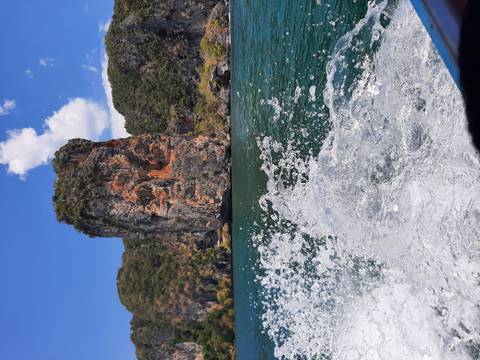       View from a speeding boat as spray rises before a massive vertical limestone karst in emerald waters.
  