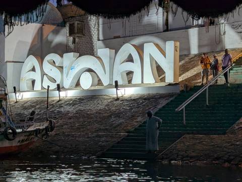       Illuminated ‘ASWAN’ sign on riverside steps at night with tourists posing nearby.
  