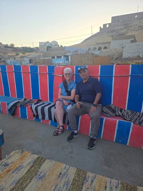       Smiling traveller and local guide seated on a colourful Nubian bench in the Nile village.
  