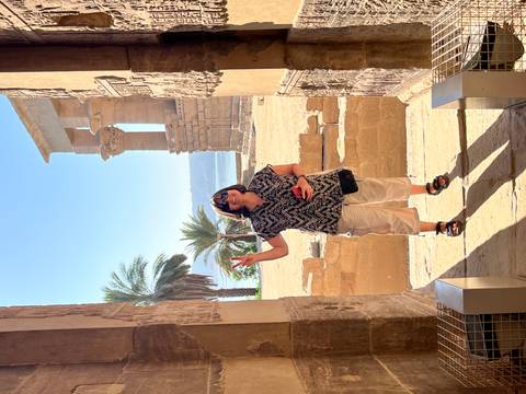       Woman flashes a peace sign inside Philae Temple with palm trees and Nile behind her.
  