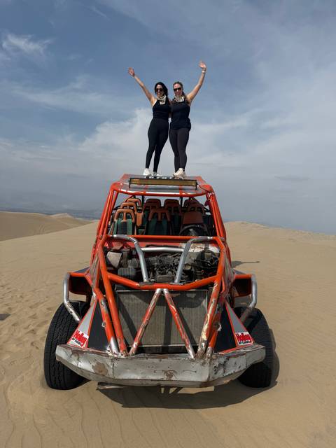       Two people stand on the roof rack of an orange dune buggy amidst vast desert sand dunes.
  