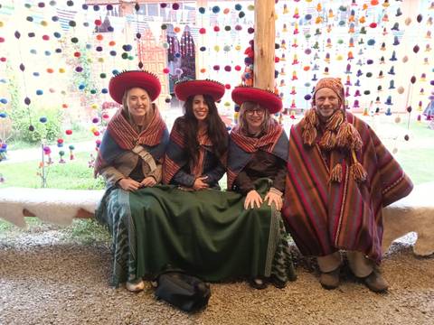       Travellers dressed in colourful traditional Quechua garments sit smiling inside a textile workshop.
  