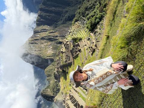       Traveller kneels smiling with the iconic ruins of Machu Picchu and Huayna Picchu peak shrouded in mist behind.
  