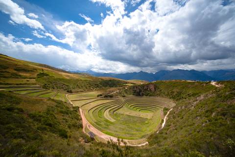       Circular agricultural terraces of Moray set amid green Andean hills beneath dramatic cloud formations.
  