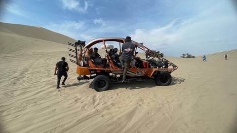       Orange dune buggy with travellers prepares to drive across vast desert sand dunes under blue sky.
  