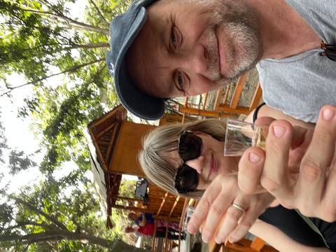       Smiling couple holding up shot glasses for a toast at an outdoor venue surrounded by trees.
  