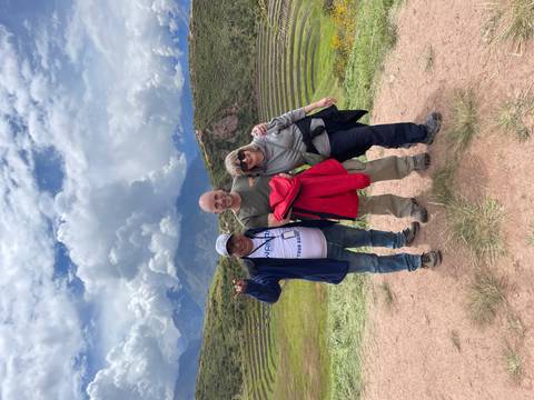       Three travellers cheerfully pose before terraced Incan ruins and dramatic cloud-filled Andean backdrop.
  