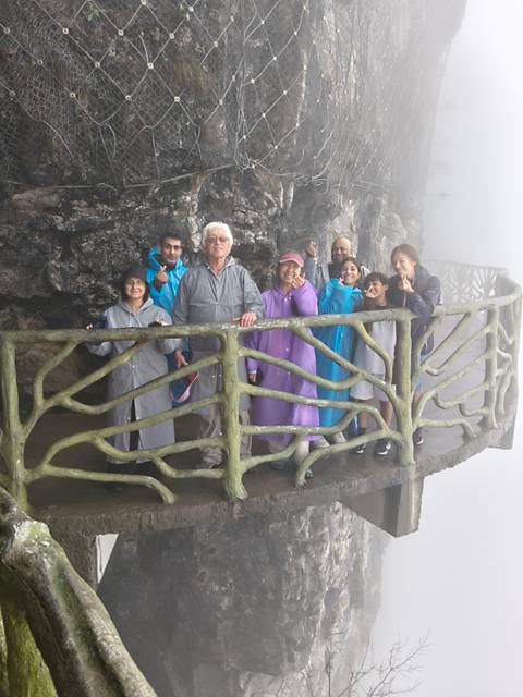       Smiling multi-generational group in colorful rain ponchos posing on a carved stone balcony in the mist.
  