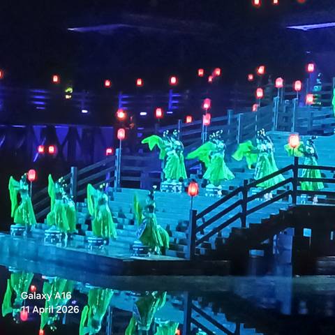       Nighttime stage performance with dancers in vivid green costumes descending illuminated steps amid red lanterns.
  