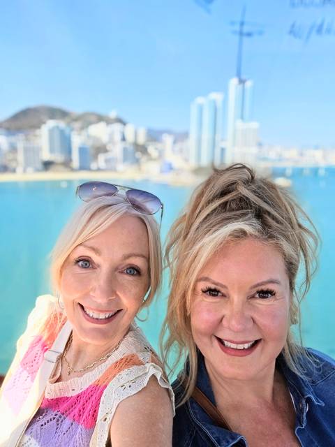       Close-up selfie of two smiling women with turquoise bay water and Busan city skyline blurred in the background.
  
