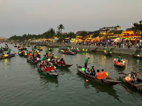       Dozens of small boats with colorful lanterns and life-jacketed passengers celebrating on a riverside at dusk.
  