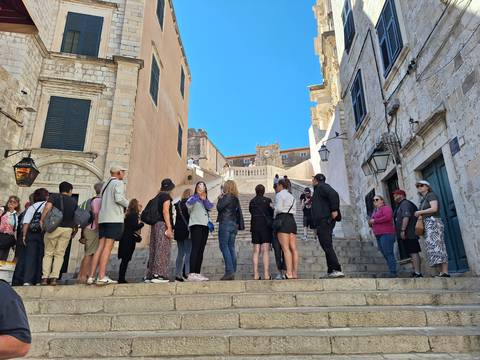       Tourists queue on stone steps between historic limestone buildings under a clear sky.
  