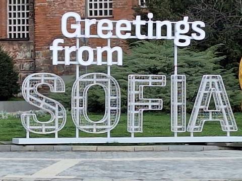       White ‘Greetings from SOFIA’ installation in front of greenery and brick façade.
  