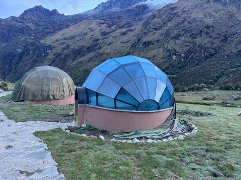       Futuristic blue-framed dome tents set on a grassy mountain campsite with steep green slopes behind.
  