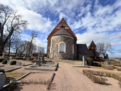       Medieval stone church with patterned brick roof surrounded by old gravestones under patchy clouds
  
