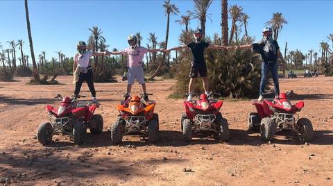       Four riders stand on red and orange quad bikes in an arid palm grove, arms outstretched in celebration.
  