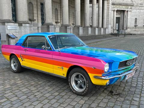       Vintage Ford Mustang painted in bright rainbow stripes parked on cobblestone plaza beside grand columns.
  