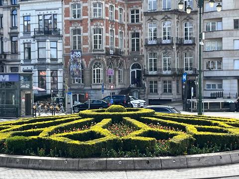       Geometric flowerbed and trimmed hedges adorn a city square lined with ornate European townhouses.
  