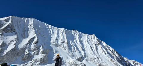       Trekker bundled in winter gear stands below a towering icy Himalayan ridge.
  