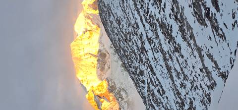       Golden sunlight ignites a snow-clad mountain summit above a rocky, snowy slope.
  