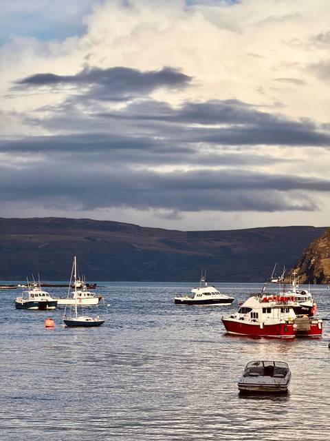       Small boats float on a calm Scottish harbor beneath brooding clouds and dark hills.
  