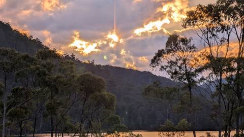       Fiery sunset breaks through clouds above a forested Tasmanian lake.
  