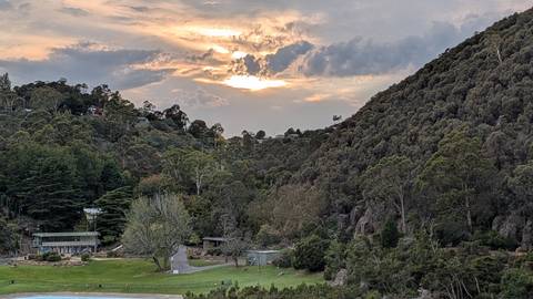       Soft sunset light filters over wooded hills and a valley homestead in Tasmania.
  